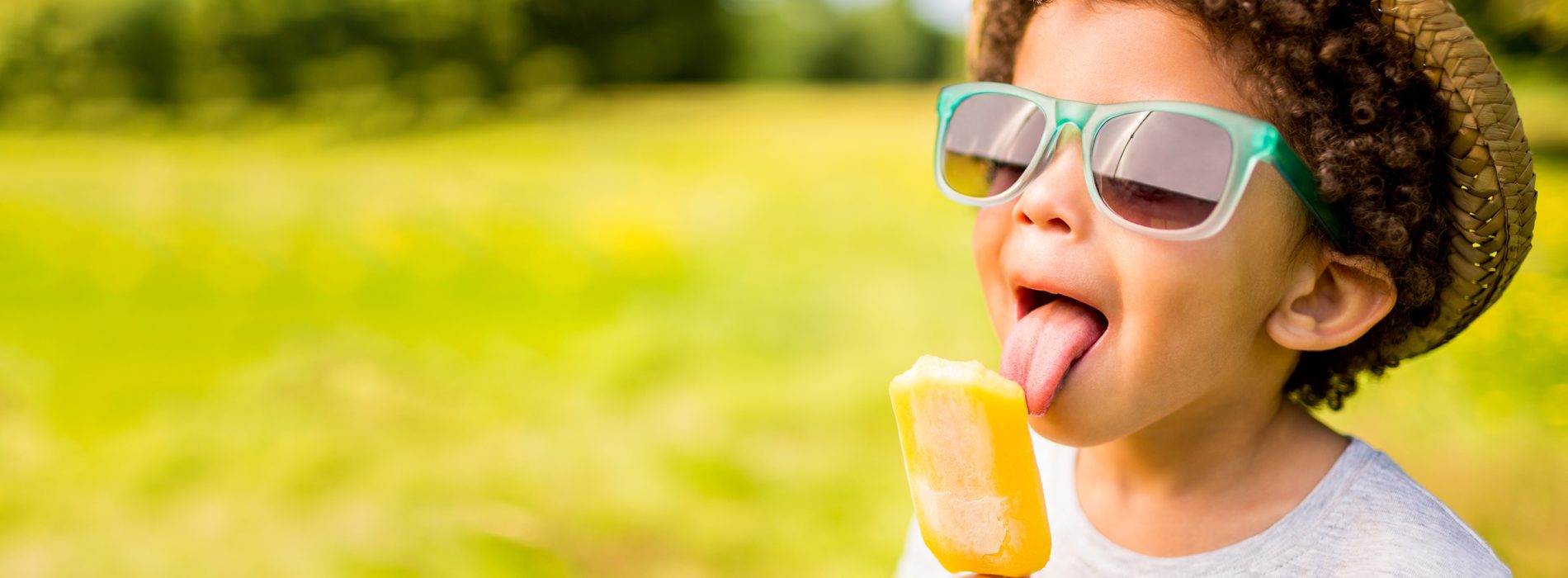 The tongue of a young boy with curly hair is touching an orange frozen treat.  He is wearing a gray t-shirt, a straw hat and blue rimmed glasses.  The background is blurry and consists of an open field of green grass with trees and bushes in the distance.